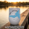 A white travel tumbler featuring a blue outline of Lake Gregory rests on a wooden dock at sunset.