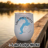 A white travel tumbler with a blue map of Lake Pend Oreille sits on a wooden dock at sunset.