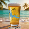 Glass of beer with "TINY LAKE" and compass design, on beach with palm trees.