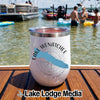 A white insulated tumbler with Lake Wenatchee printed on it sits on a wooden dock overlooking a lake with boats and swimmers.