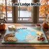 A glass cutting board displaying a map of Floyd Lake, Minnesota, surrounded by crackers, cheese, and pecans on a wooden table.