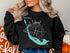 Black crewneck sweatshirt with a teal compass and Sanibel Island, Florida map graphic.
