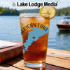 Glass of iced tea with "American Lake" graphic and lemon slice, by a lake.
