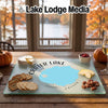 A glass cutting board featuring a map of Crater Lake rests on a wooden table with crackers, cheese, and pecans.