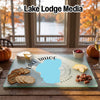 A rectangular glass cutting board featuring a blue Lake Tahoe map graphic, surrounded by crackers, cheese, and fruit on a wooden table.