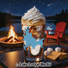 A glass of iced coffee with whipped cream sits by a campfire and lake.