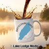 Dark liquid is poured into a white ceramic mug featuring a Rattlesnake Lake graphic, outdoors against a misty lake background.