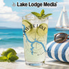 Clear glass with drink, lime, and mint on beach with sailboat and sunglasses.