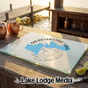 A rectangular glass cutting board rests on a wooden table, featuring a blue map of Merrymeeting Lake, New Hampshire, over a compass rose design.