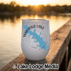 A white travel mug with a blue map of Waurika Lake rests on a wooden dock overlooking a lake at sunset.