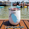 A white insulated wine tumbler with a Black Lake New York graphic rests on a wooden boat deck, with people swimming in the lake behind it.