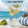 Glass with drink, limes, mint, and sunglasses on beach with sailboat.