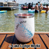 A white travel tumbler with a Lake Anna compass design sits on a wooden boat deck overlooking people in a lake.