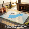 A glass cutting board featuring a blue map graphic of Convict Lake, California rests on an outdoor wooden table near copper mugs and a dock.