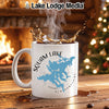 White mug with Squam Lake map and compass design, steaming coffee, fireplace, Christmas tree.