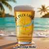 Glass of beer with "Port Peck Lake" text and compass on a beach with palm trees.