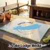 A glass cutting board displaying a lake map, compass rose, and Sand Lake Ontario text rests on an outdoor wooden table with copper mugs and lime slices.