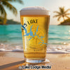 Glass of beer with "Falls Lake" text and compass on a sandy beach.