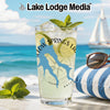 Glass with yellow drink, ice, mint, and lime on beach with sailboat.