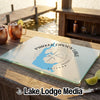 A tempered glass cutting board featuring a blue map and compass design of Parker Canyon Lake, Arizona rests on an outdoor wooden table with copper mugs nearby.