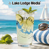 Glass with drink, limes, mint, and sunglasses on beach with sailboat.