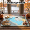 A glass cutting board printed with Sylvan Lake, South Dakota, surrounded by crackers, cheese, and pecans on a wooden table.