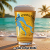 Pint glass of beer with blue lake outline and compass on a beach background.