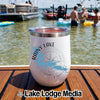 A white stainless steel wine tumbler with a Rainy Lake compass design rests on a wooden boat deck with people in a lake background.