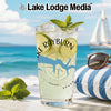 Glass with drink, lime, mint, and sunglasses on beach with sailboat.