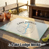 A glass cutting board featuring a Lake Barkley compass design rests on a wooden table near copper mugs.
