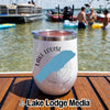 A white tumbler with a Lake Louise graphic rests on a wooden boat deck in front of people swimming in a lake.