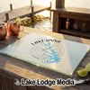A glass cutting board printed with a Lake Anna map and compass design sits on a wooden dock with copper mugs nearby.