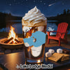 A tall glass of iced coffee topped with whipped cream, sitting on a table outdoors at night near a campfire and a lake.
