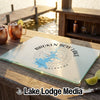 A tempered glass cutting board featuring a compass design and the text Broken Bow Lake Oklahoma rests on a wooden bar overlooking a lake.