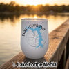 A white insulated wine tumbler featuring a blue lake map and compass design rests on a wooden dock at sunset.