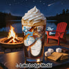 A tall iced beverage topped with whipped cream sits on a table outdoors next to s’more ingredients, with a campfire and lake in the background.