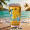 Pint glass with Medina Lake outline, compass, and text on beach with ocean background.