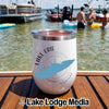 A white insulated wine tumbler with a Lake Erie graphic sits on a wooden deck by a lake with people swimming.