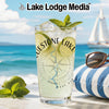 A tall glass filled with a refreshing iced drink, garnished with lime and mint, set against a lakeside backdrop.
