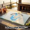 A rectangular glass cutting board with a map of Broadway Lake, South Carolina rests on a wooden bar overlooking a lake.