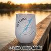 A white stemless tumbler with a blue map of Key Largo, Florida, and a compass rose graphic sits on a wooden dock at sunset.