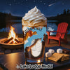 A glass with a layered drink sits on a table, with a campfire and lake scene in the background.