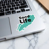 Sticker with "Sand Life" text on a teal lake outline on a laptop.