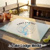A rectangular glass cutting board printed with a compass design and Lake Oconee, Georgia, rests on a wooden dock beside two copper mugs.
