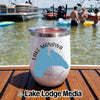 A white insulated tumbler with a Lake Manawa design rests on a wooden dock with boats and people swimming in the background.