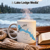 White mug with "Bass Lake California" design and steaming coffee in front of a lake view.