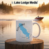 White mug with Gull Lake Michigan map and compass on a dock with a lake background.