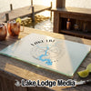 A glass cutting board featuring a map of Lake LBJ and a compass rose rests on an outdoor wooden table with copper mugs.