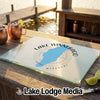 A glass cutting board featuring a blue map of Lake Winnebago, Missouri, and compass rose design rests on a wooden bar overlooking a lake.