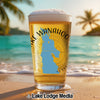 A pint glass filled with beer stands on a sunny beach, featuring a Lake Wanahoo Nebraska map design.